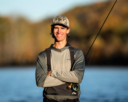 Cam looking and smiling at the camera, with arms crossed while holding onto his fly rod