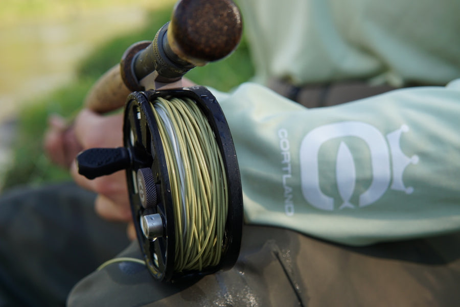 Closeup shot of the bottom of the fly rod, which has Long Belly Distance fly line. The coil is moss green and white. Someone is holding the rod on their lap.