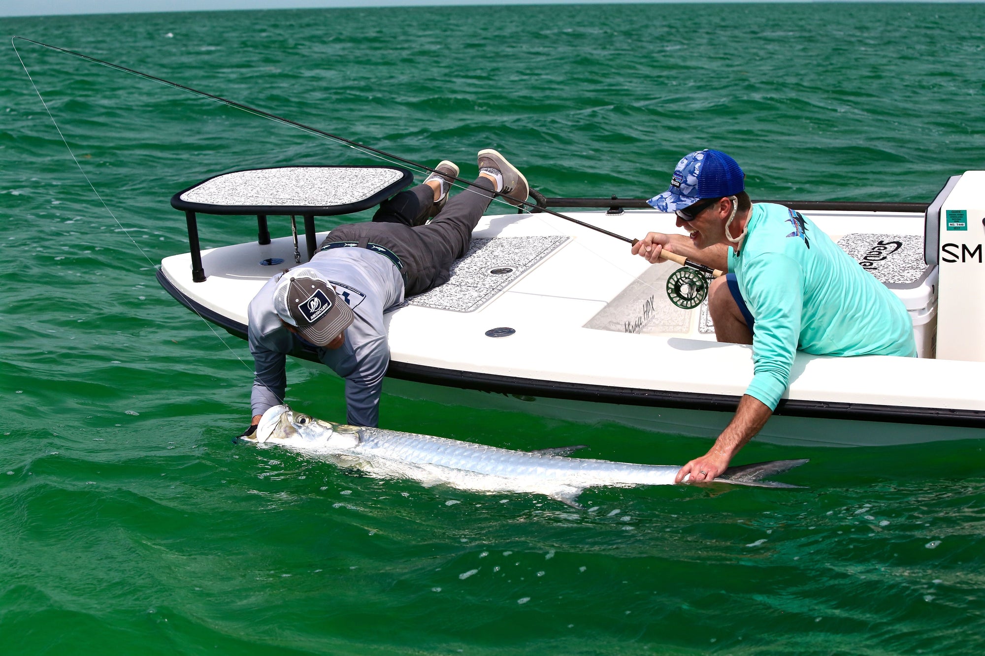 Two individuals are leaning over a boat and holding onto a tarpon. 